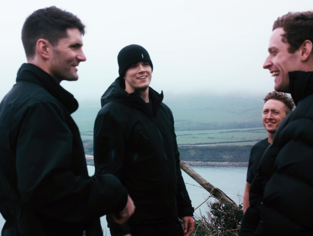 Four people in outdoor jackets stand together, talking beside a viewpoint overlooking water and hills.