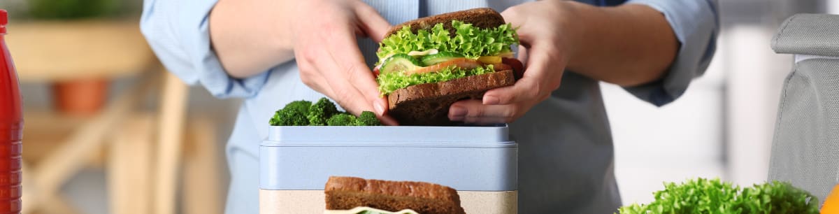 A picture of someone preparing their packed lunch, the person is holding a salad sandwich on brown bread.