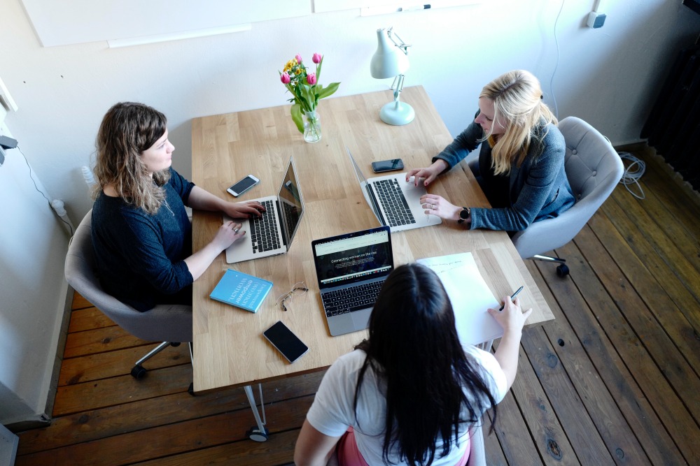Three people sit around a table using laptops and reviewing documents during a meeting.