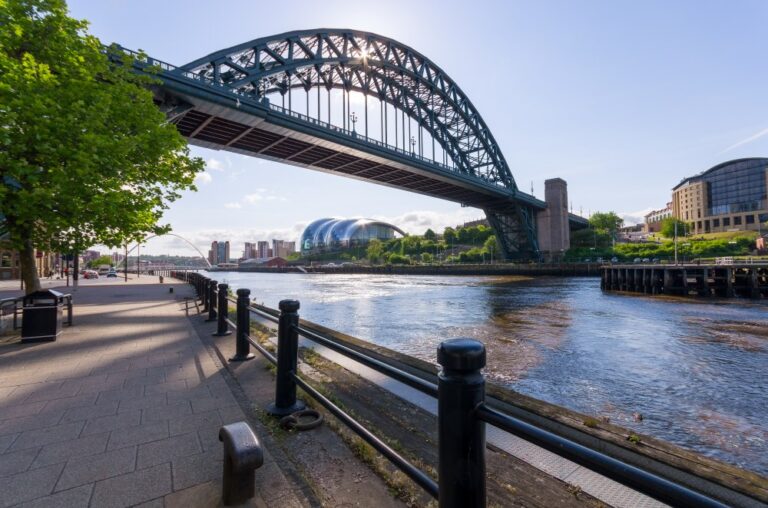 Arch bridge spans a river beside a walkway with railings and buildings along the waterfront.