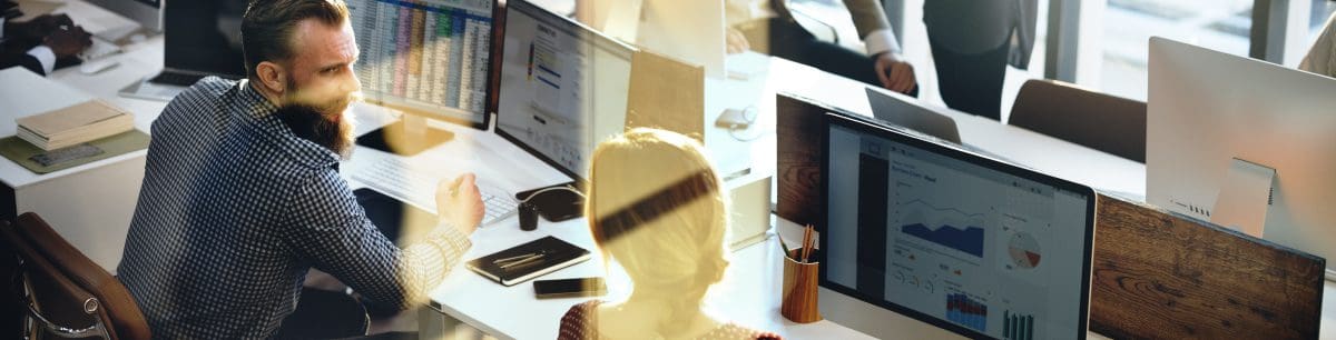 2 colleagues sat side by side at a desk, view from behind so their computer screens are visible