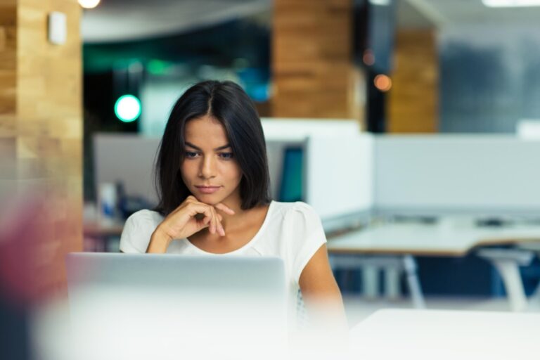 Person rests chin on hand while looking at a laptop screen in a modern office.