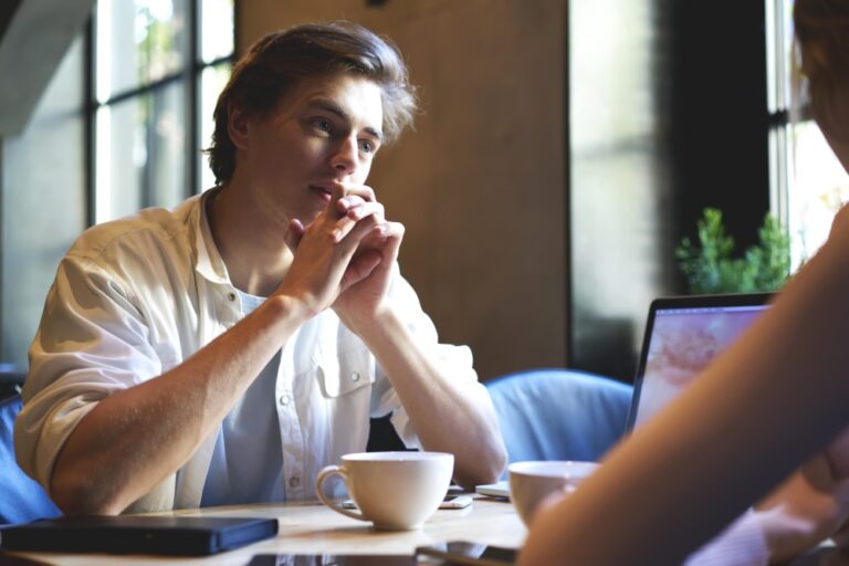 Close-up of a person seated at a table listening, with a laptop and coffee cups nearby.
