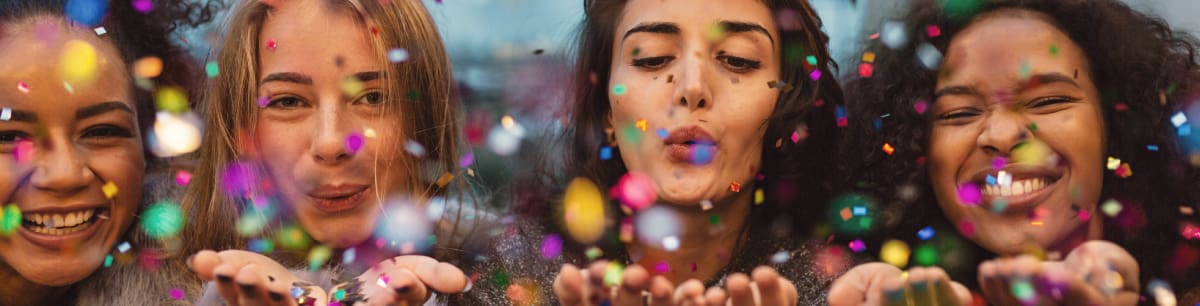 4 women all blowing confetti at the camera