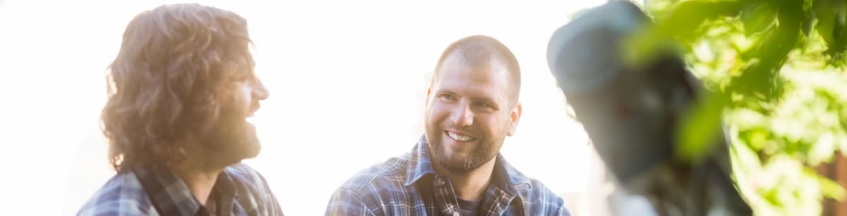 Two men sitting outdoors, talking and smiling, with a bright background.