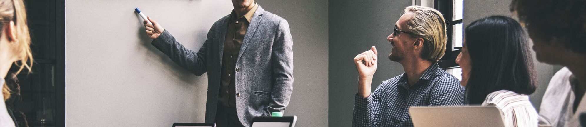 Business meeting with one man standing at the front while others sit around a table.