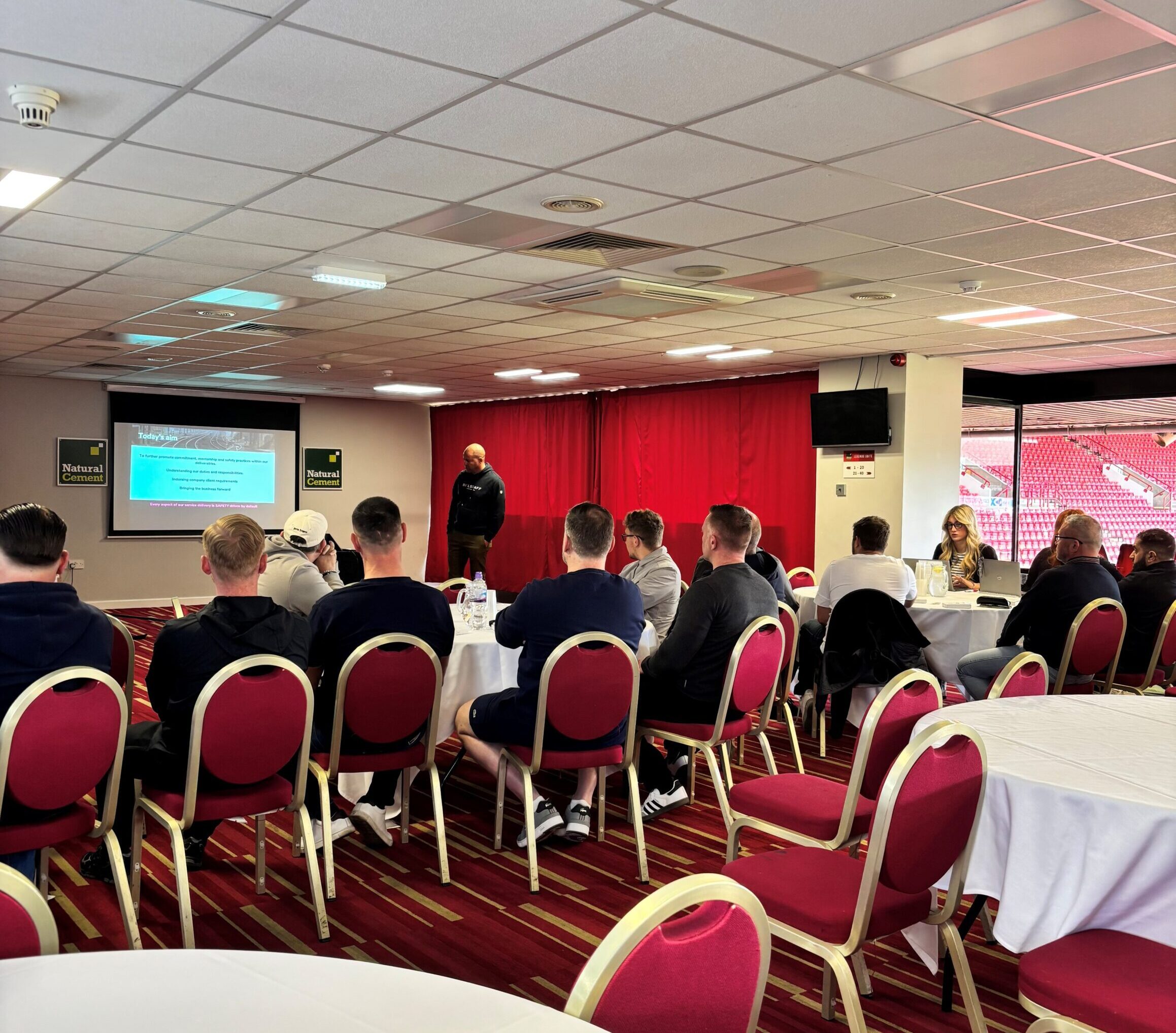 Group seated in a stadium lounge watching a safety presentation on a screen.