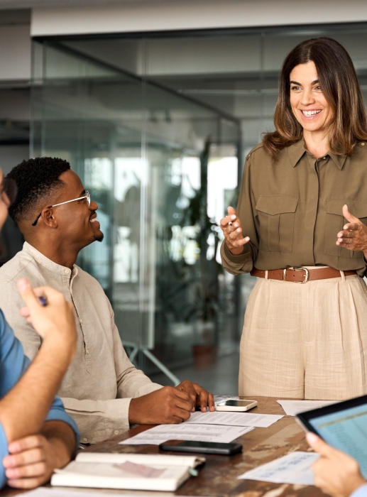 Person standing and speaking to colleagues seated at a table with documents and devices.