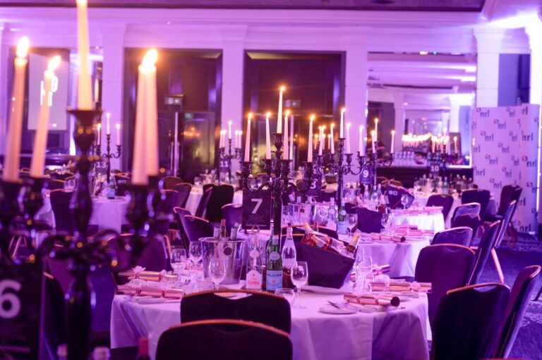 Round tables set for an event with tall candles, glassware and place settings in a lit venue.