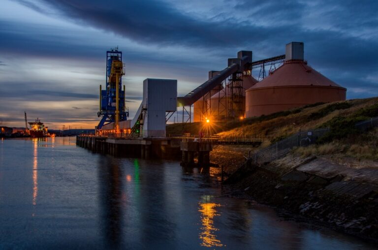 Industrial buildings and structures at Blyth dock beside water, with lights reflecting at dusk.