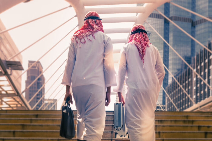 Two people in traditional clothing walk across a modern city bridge carrying briefcases.