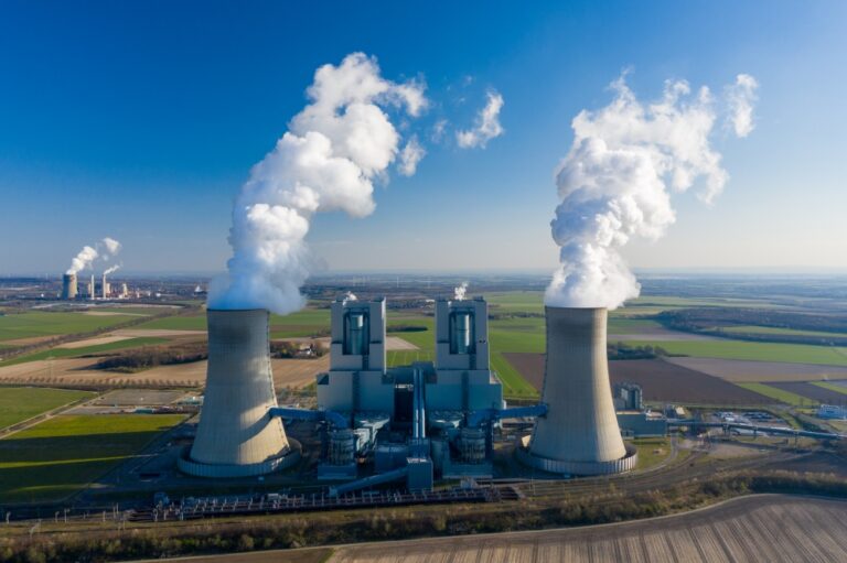 Industrial power plant with cooling towers emitting steam across farmland under a clear sky.