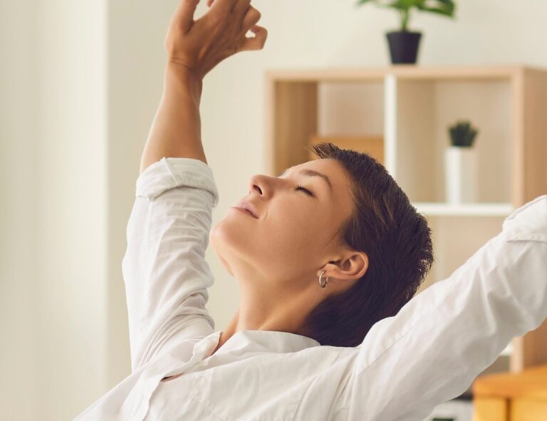 Person stretches arms upward while seated indoors with shelves and a plant in the background.