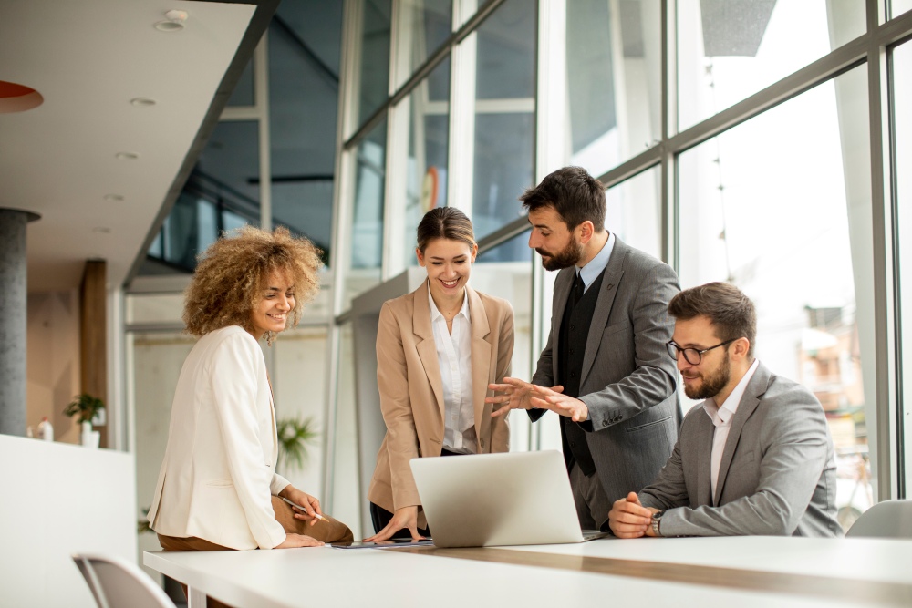 Four people stand around a table, reviewing a laptop and discussing work together.