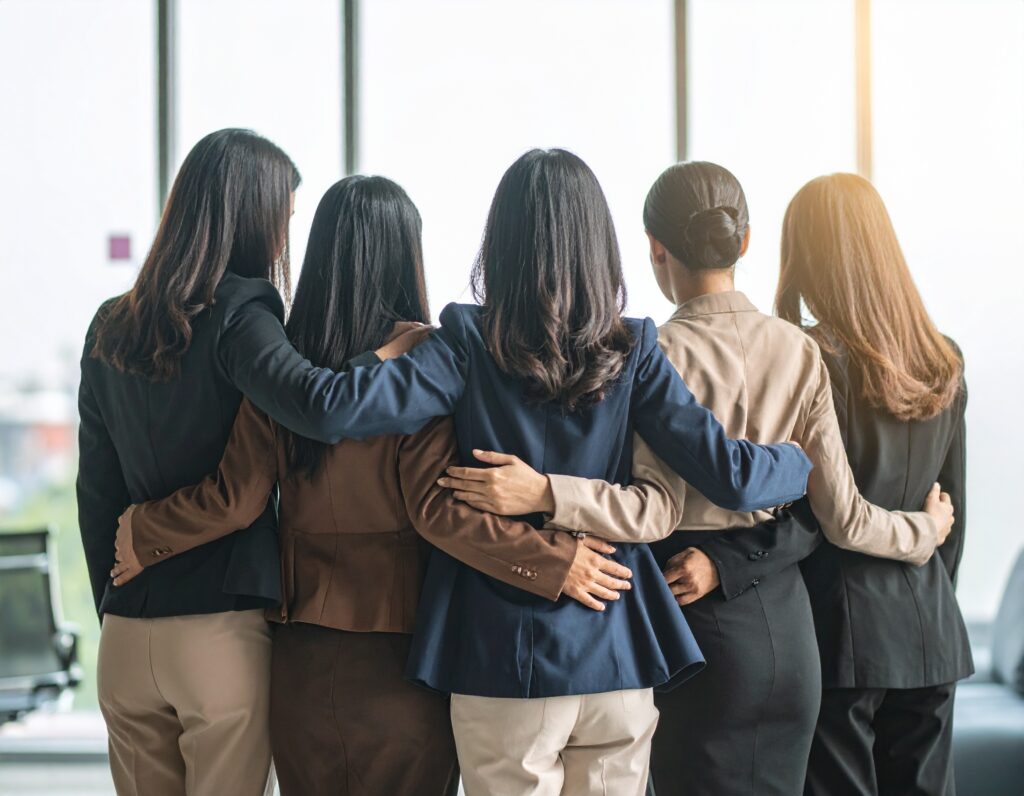 Five women stand together with arms around each other in a modern office setting.