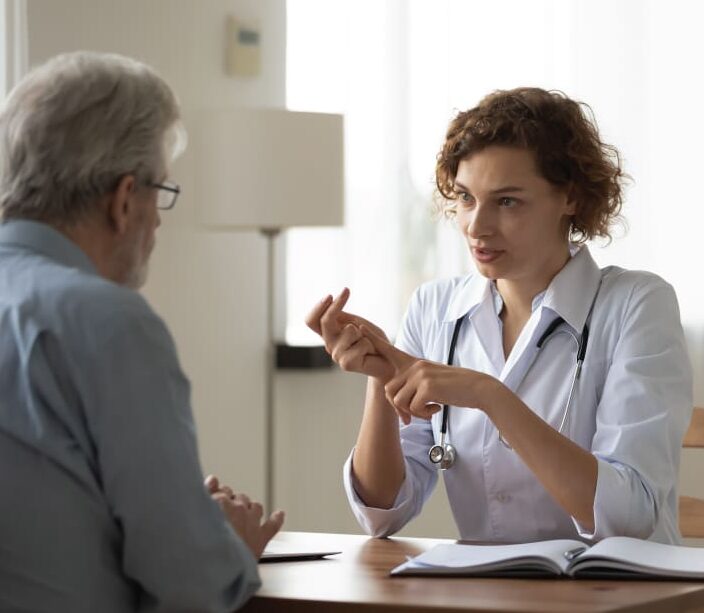 Medical professional speaks with a seated person while pointing at notes during a consultation.