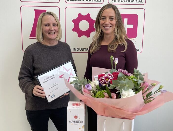 Two people stand holding a framed certificate and bouquet in front of a branded display wall.