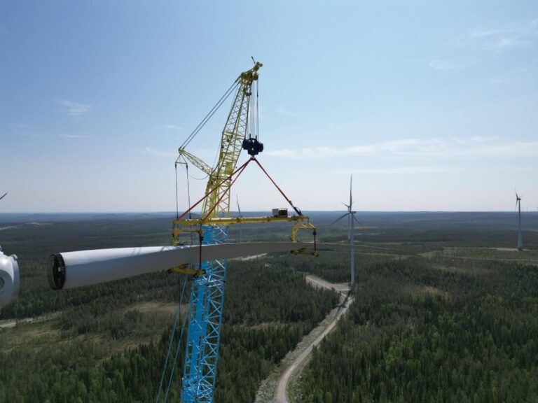 Crane lifts a wind turbine blade above a forested landscape with turbines in the distance.