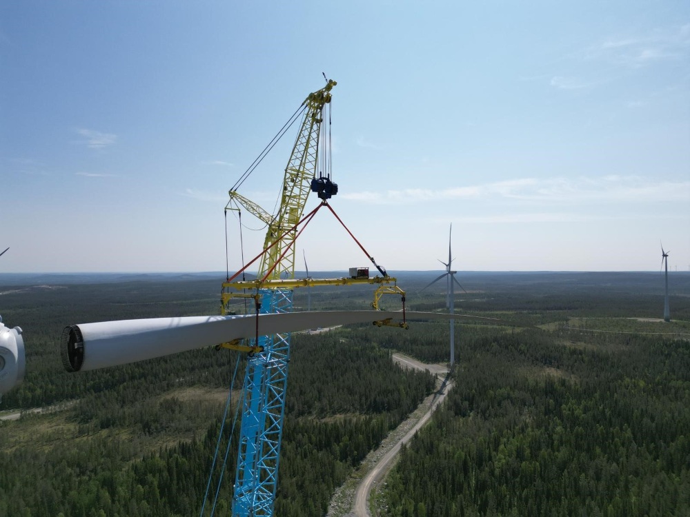 Crane lifts a wind turbine blade above a forested landscape with turbines in the distance.