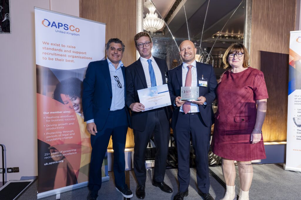 Four people stand together holding an award and certificate beside an APSCo branded display.