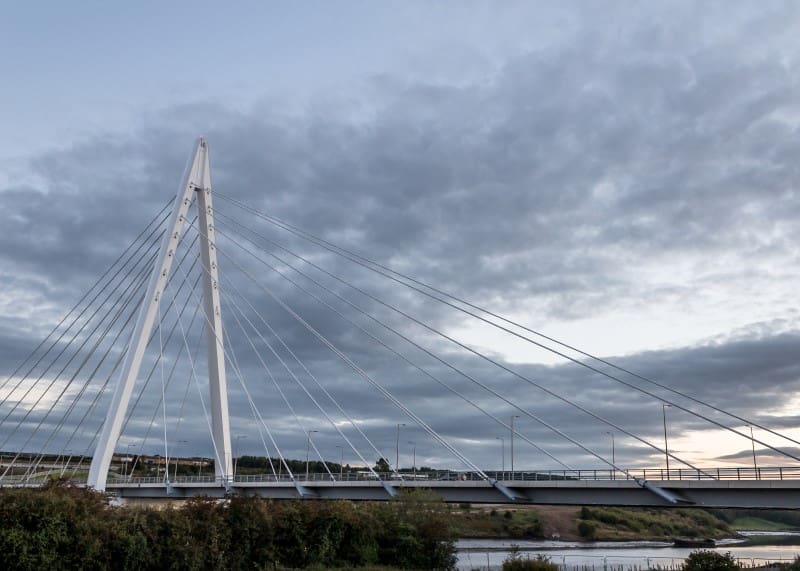 Bridge with tall central mast and angled cables spanning a river under a cloudy sky.