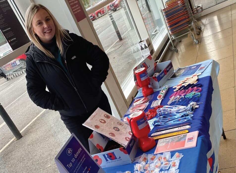 Person stands beside a table displaying poppies, donation items and information materials indoors.