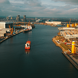 Aerial view of a cargo ship sailing through a canal lined with industrial buildings and cranes.