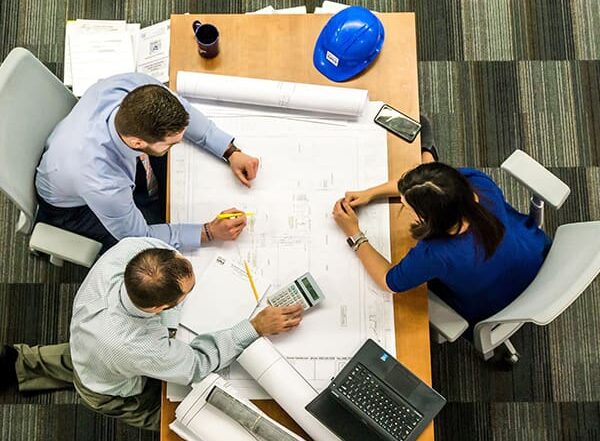 Three people sit around a table reviewing large technical drawings with a laptop and tools.