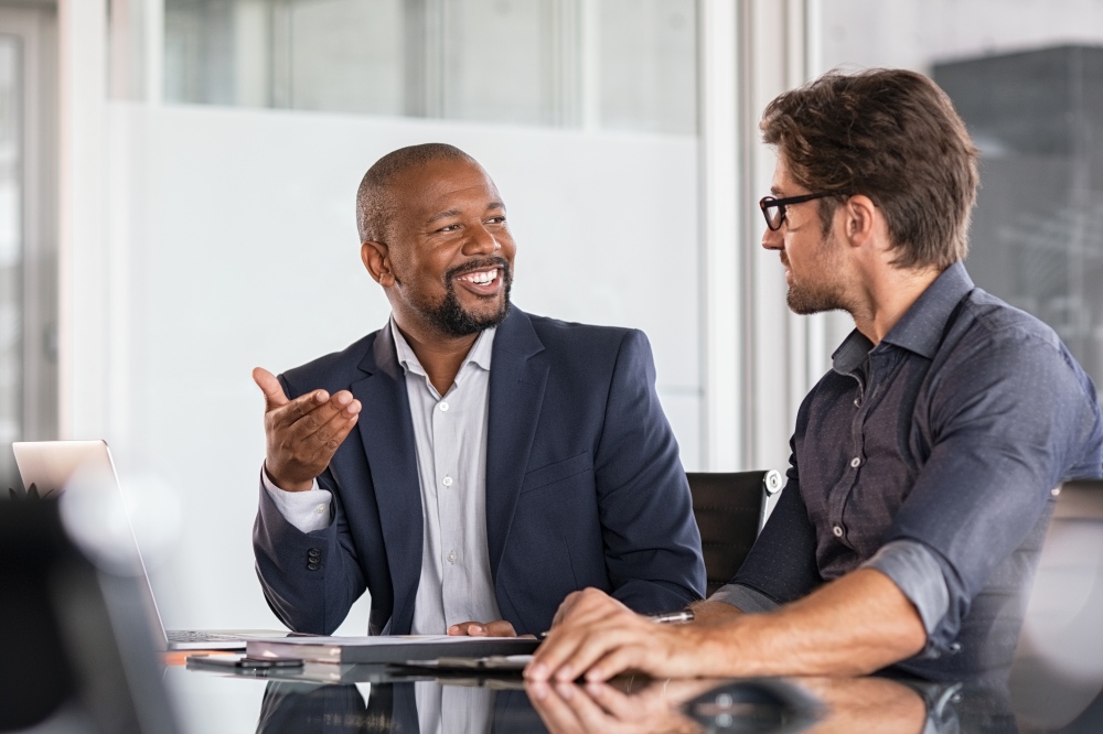 Two people sit at a desk, one gesturing while both review work during a discussion.