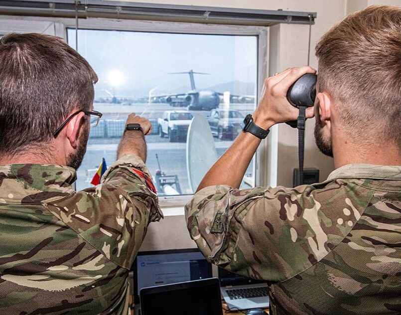 Two people in military uniform look through a window using binoculars towards aircraft outside.
