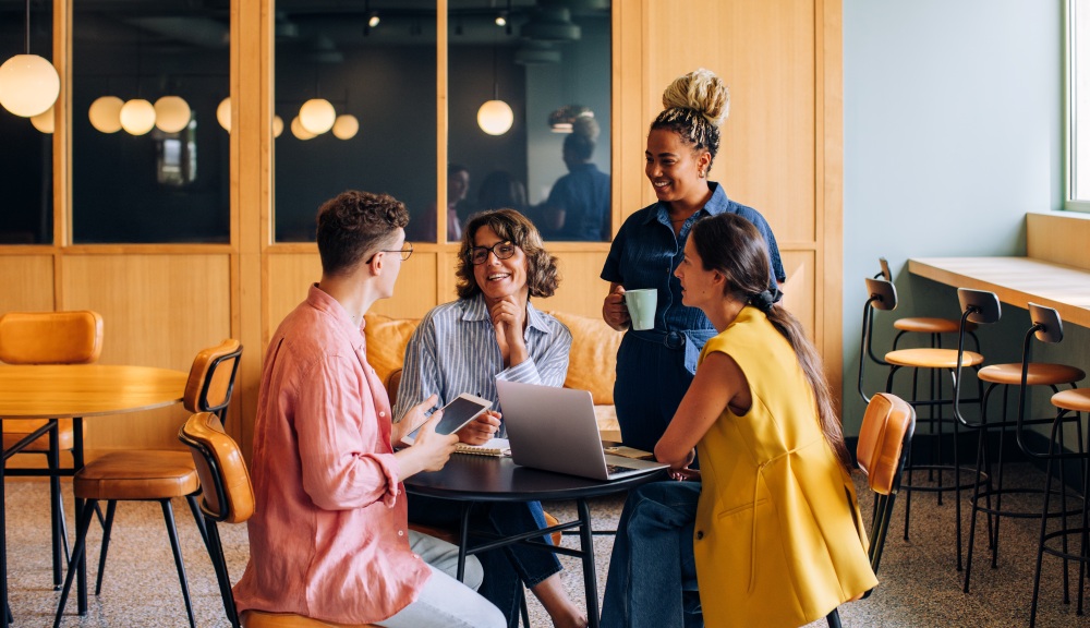 Four people gather around a table with a laptop, talking and reviewing work in a modern office.