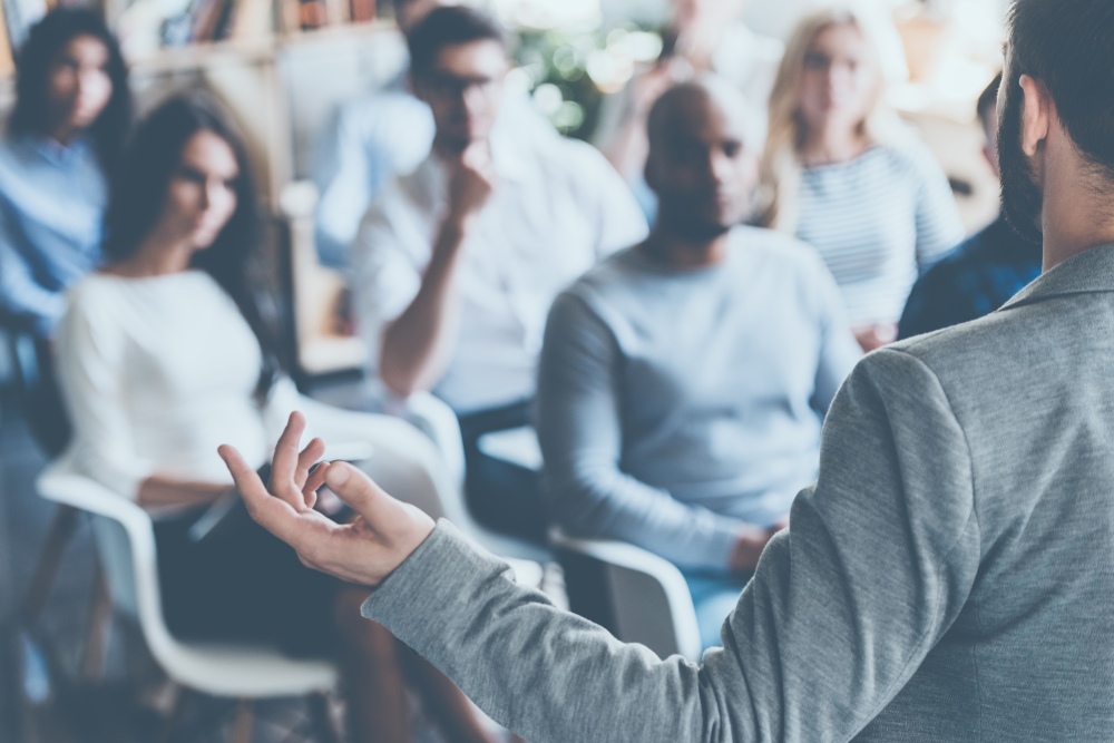 Person stands speaking to a seated group while gesturing during a presentation.