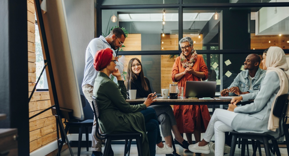 Six people sit and stand around a table, talking and working together in a glass-walled office.