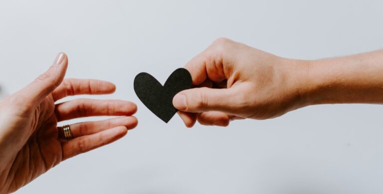 One hand passes a small heart shape to another hand against a plain background.