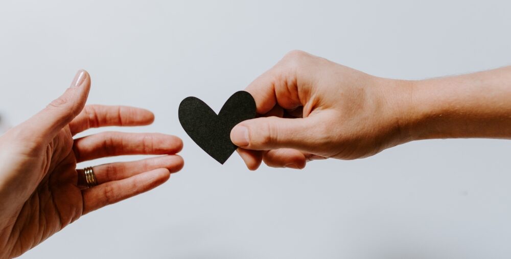 One hand passes a small heart shape to another hand against a plain background.