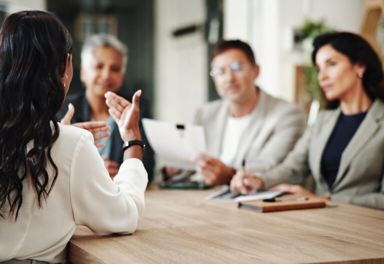 Person speaks while gesturing across a table as others listen and review notes in a meeting.