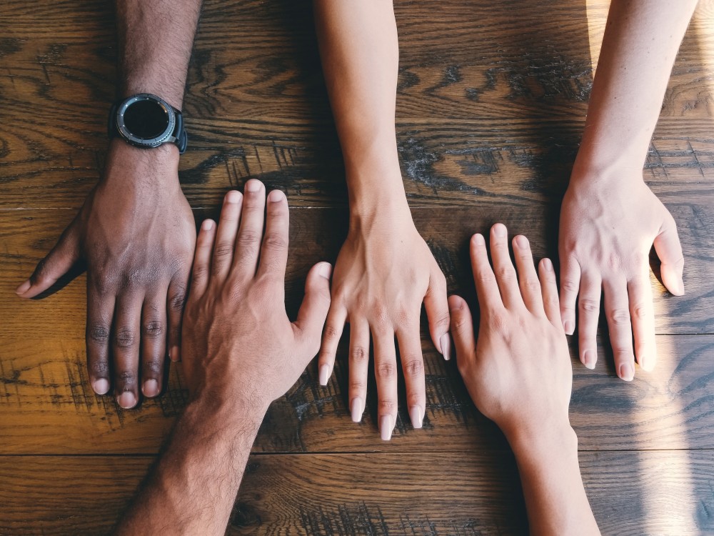 Four hands placed side by side on a wooden table, showing different skin tones.