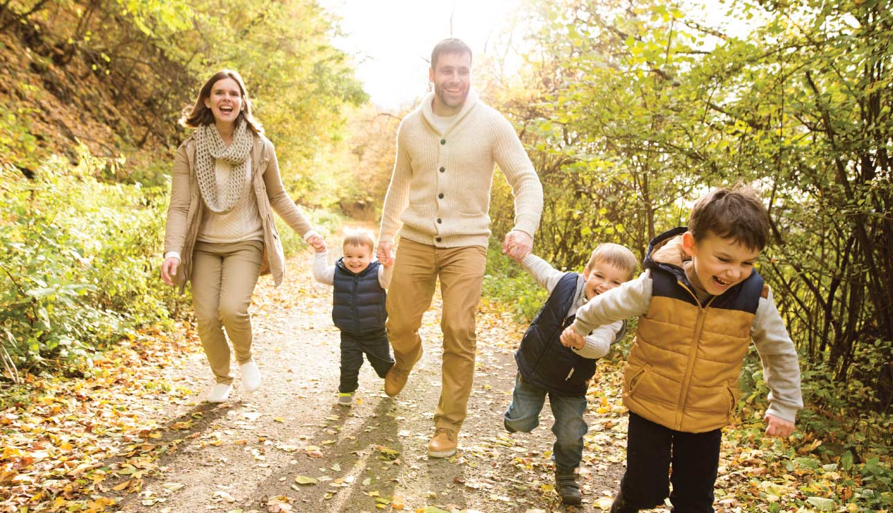 Four people walk along a wooded path, with two holding hands with children in autumn sunlight.