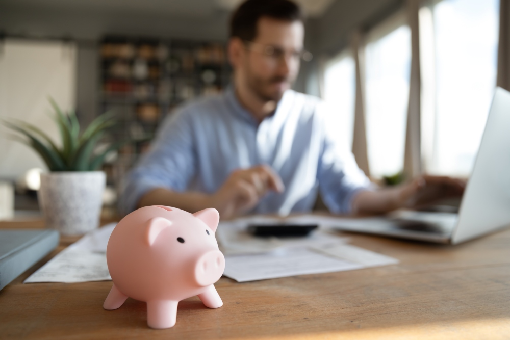 Close-up of a piggy bank on a table with a person using a laptop and reviewing papers in the background.