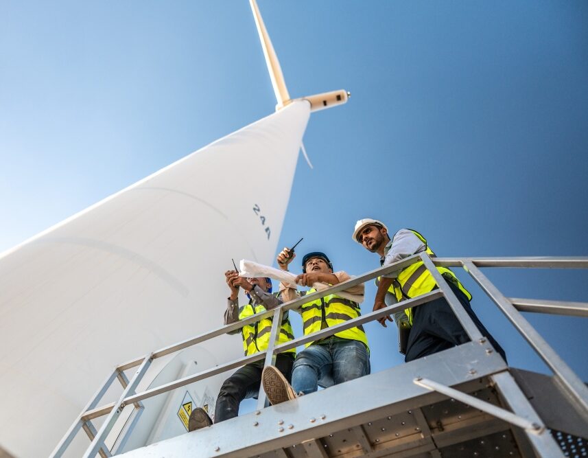 Three people in safety helmets and high-visibility vests stand on a platform near a wind turbine.