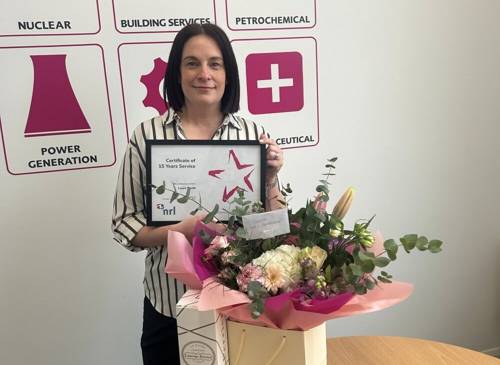 Person holds a certificate and bouquet while standing in front of a wall with industry icons.