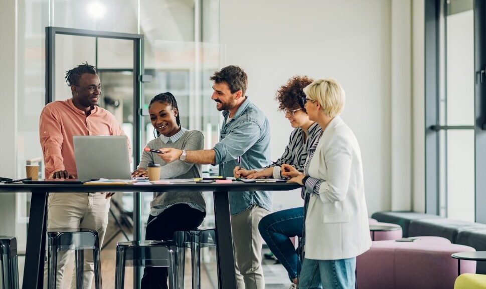 Five people stand and sit around a high table, discussing work with a laptop and notes indoors.