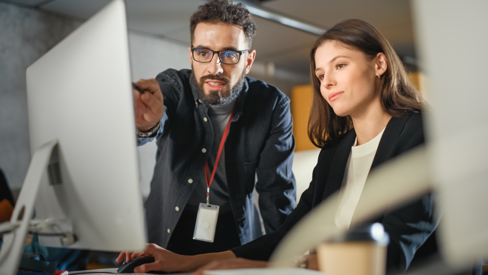 Two people sit at a computer, one pointing at the screen while discussing work.