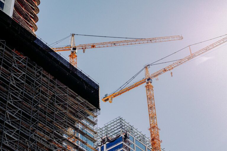Two tower cranes stand beside a high-rise construction building with scaffolding and framework.