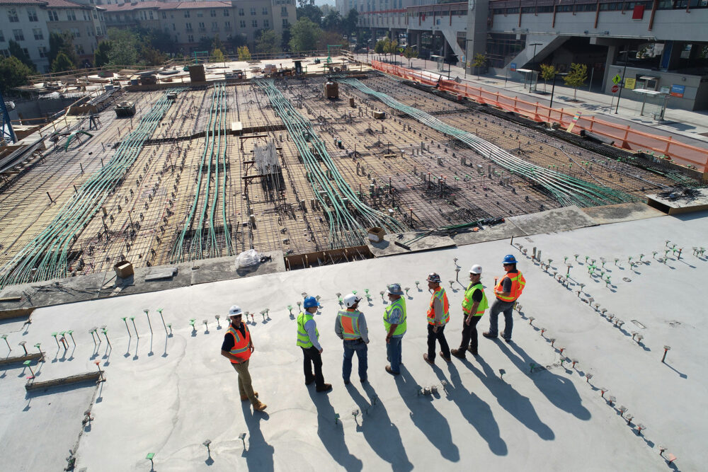Seven workers in safety helmets stand on a concrete slab overlooking a large construction site.