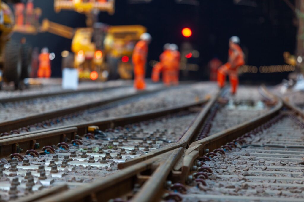 Railway workers in high-visibility clothing stand on tracks during maintenance with equipment nearby.