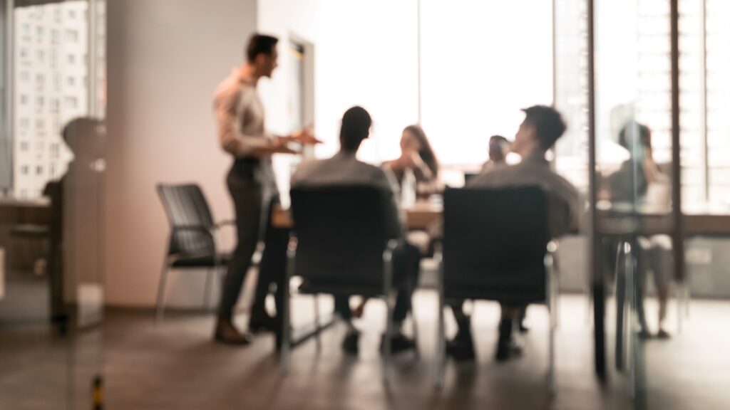 Blurred view of a person presenting to a seated group in a meeting room with large windows.