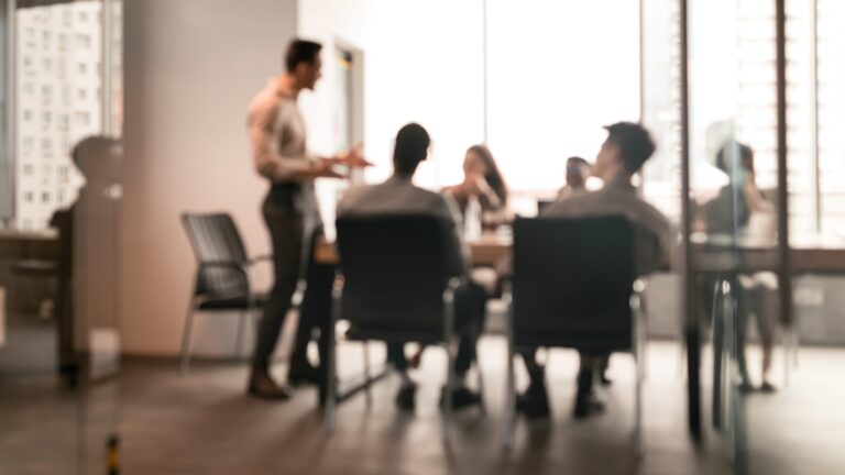 Blurred view of a person presenting to a seated group in a meeting room with large windows.