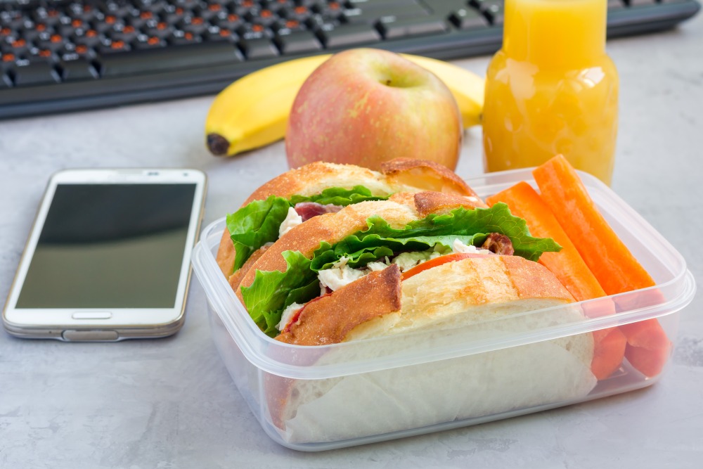 Sandwich, apple, banana, carrot sticks and juice in a container beside a phone and keyboard.