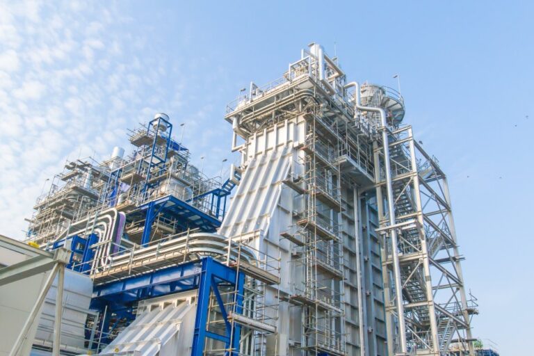 Large industrial facility with steel framework, pipes and platforms against a clear blue sky.